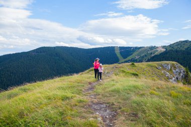 Mother and daughter with backpack hike on mountain trail, beautiful summer day, mountain nature landscape. Family outdoors activity.