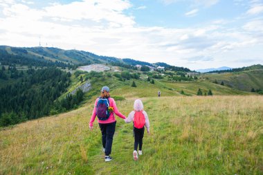 Mother and daughter with backpack hike on mountain trail, beautiful summer day, mountain nature landscape. Family outdoors activity.