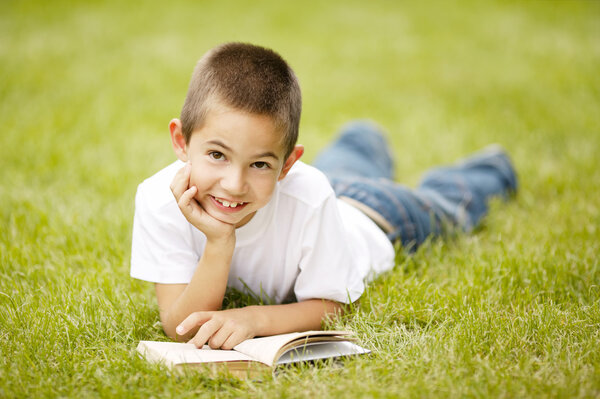 Маленький счастливый мальчик читает книгу, лежащую на траве
