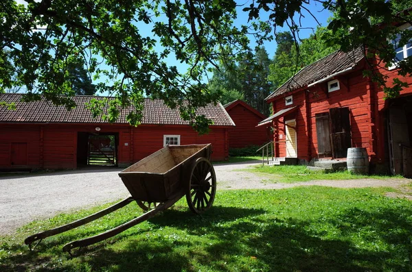 2019年7月3日 瑞典瓦尔比露天博物馆 Vallby Open Air Museum 的瑞典传统红房子环绕的院子里绿色草地上的大轮子木车 夏日晴天 — 图库照片