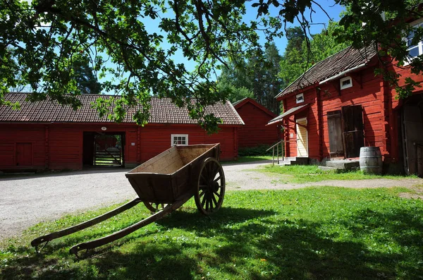 2019年7月3日 瑞典瓦尔比露天博物馆 Vallby Open Air Museum 的瑞典传统红房子环绕的院子里绿色草地上的大轮子木车 夏日晴天 — 图库照片