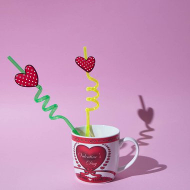 İki eğri pipet ve pembe arka planda gölgesi olan bir aşk bardağında kalpler. Minimalist aşk sahnesi.