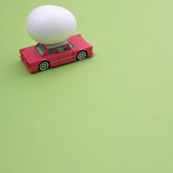 The Easter white egg is transported by an old pink car on a light green surface. Minimalistic scene.