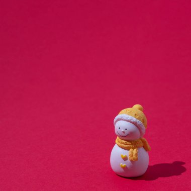 Sarı eşarplı ve şapkalı bir kardan adam kırmızı arka planda duruyor. Minimalist kış sahnesi.