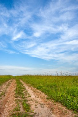 Tarla ve toprak yol, ufkun ötesine gidiyor, bulutlu mavi gökyüzünün arka planına doğru..