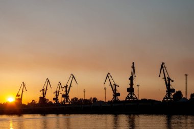 Gün batımındaki kızıl gökyüzünün arka planına karşı kule vinçlerinin siluetleri.