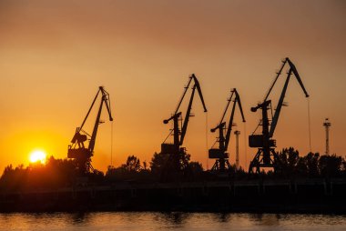 Gün batımındaki kızıl gökyüzünün arka planına karşı kule vinçlerinin siluetleri.