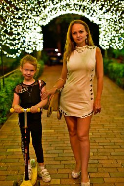 Bir çocuklu güzel bir kadın, bir kız, akşam parkında yürüyor..