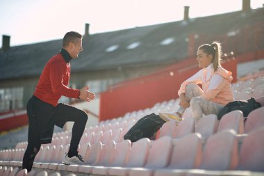 Kafkasyalı kişisel antrenör konuşuyor, anlatıyor, genç yetişkin bir kadına gülümseyerek ve el hareketleriyle. 
