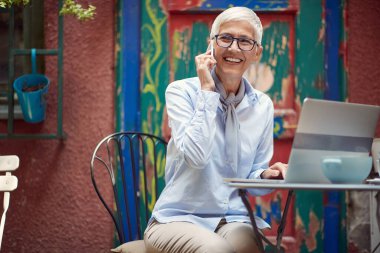 Kafkasyalı yaşlı güzel bir kadın açık kafedeki masada oturuyor, cep telefonuyla konuşuyor, gülümsüyor.