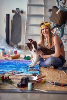 Seksi ve çekici bir kadın sanatçı stüdyoda rahat bir atmosferde yerde oturuyor ve resim üzerinde çalışırken köpeğiyle fotoğraf çektirmek için poz veriyor. Sanat, resim, stüdyo