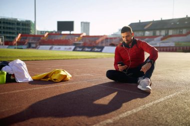 Genç beyaz sakallı adam spor giyim stadyumunda atletizm pistinde oturuyor ve cep telefonuna bakıyor.