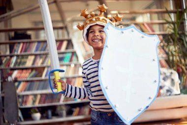 Şövalye gibi giyinmiş neşeli küçük bir çocuk evde rahat bir atmosferde oyun oynarken fotoğraf için poz veriyor. Aile, ev, oyun zamanı