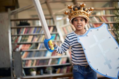 Kılıcı ve kalkanı olan neşeli küçük bir çocuk evde rahat bir atmosferde oynarken fotoğraf için poz veriyor. Aile, ev, oyun zamanı