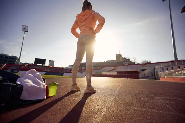 visualizing goal concept. low angle image from behind of caucasian young female standing at the athletic track with hands on hips, preparing for training,. copy space