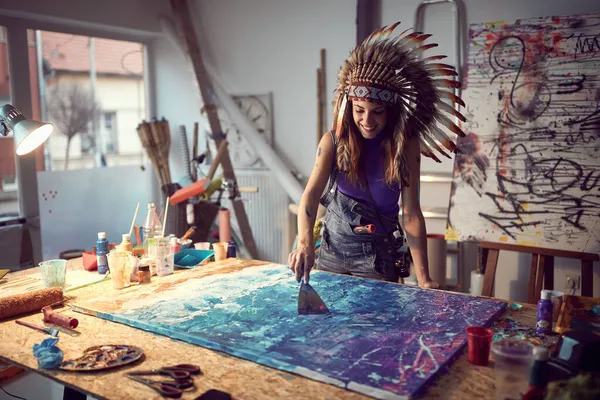 一位戴着战争帽子的年轻女画家正在工作室里愉快的气氛中 用水枪在她的新画上涂上颜料 工作室 — 图库照片