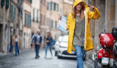 Sarı yağmurluklu genç bir kız öğrenci bulutlu bir günde müziği dinlemekten ve şehirde yürümekten zevk alıyor. Yürü, yağmur, şehir