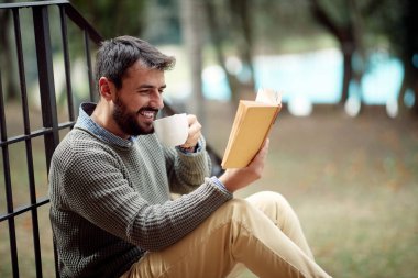 Genç yakışıklı bir adam rahat bir atmosferde evin önünde merdivenlerde otururken kitap ve kahvenin tadını çıkarıyor. Şehir, dışarısı, dinlenme
