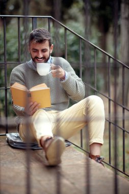 Genç yakışıklı bir adam evin önünde rahat bir atmosferde merdivenlerde oturup kitap okuyup kahve içerken iyi vakit geçiriyor. Şehir, dışarısı, dinlenme