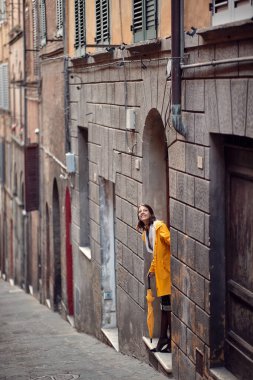 Sarı yağmurluklu genç bir kız binanın girişinde duruyor ve yağmur yağıp yağmadığını kontrol ediyor. Yürü, yağmur, şehir