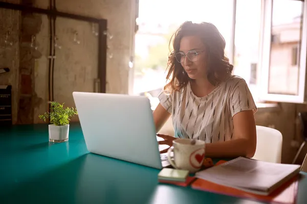 オフィスの快適な雰囲気の中で机の上に座っている若い女性がノートパソコンで働いています — ストック写真