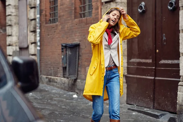 一个穿着黄色雨衣的年轻女孩在一个阴郁的雨天在城市里散步时正在打电话 — 图库照片