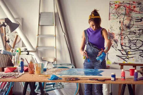 A young female artist is pouring the paint over canvas in a pleasant atmosphere in her studio. Art, painting, studio