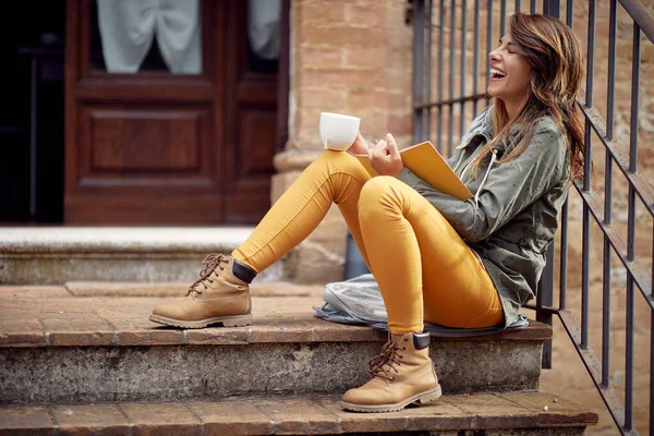 一个年轻的女孩坐在大楼前面的门廊上 在轻松的气氛中享受着一本书和一杯咖啡 — 图库照片