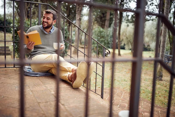 美しい日に温かいお茶を楽しみながら 若い男がポーチに座って本を読んでいます — ストック写真