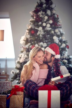 Bir adam Noel hediyesi aldığı için sevgilisini öpüyor. Noel tatili sırasında evde birlikte şenlikli bir ortamda yerde oturuyorlar. Noel, ev, ilişki, hediyeler