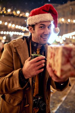 Noel Baba şapkalı, kışın açık havada gülümseyen ve Noel hediyesi veren genç adam..