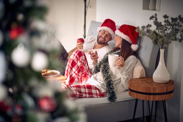 クリスマスの帽子をかぶった若いカップルは クリスマス休暇中の美しい朝にベッドで朝食を取っています クリスマス プレゼント — ストック写真