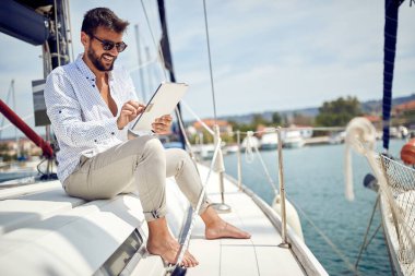 Yatta oturmuş, deniz kenarında güzel bir günde rıhtımda ata binerken tabletin keyfini çıkaran genç bir adam. Yaz, deniz, tatil