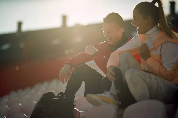身着运动服坐在体育场讲台上大声笑着的一对年轻的高加索成年人夫妇 — 图库照片