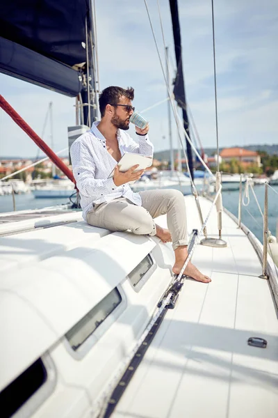 タブレットを持った若い男がヨットに乗って海辺の美しい日にドックを通っている間にコーヒーを楽しんでいます — ストック写真