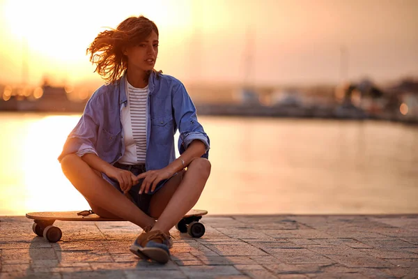 若い美しい女の子がドックのスケートボードに座って 海辺の美しい日に風の強い夕日を楽しんでいます — ストック写真