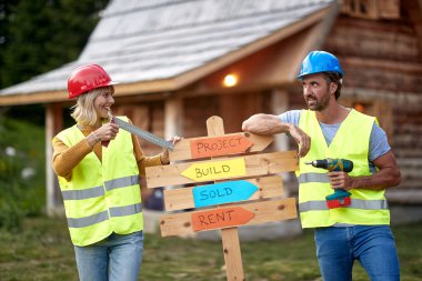 Tahtası olan mutlu, güçlü, genç bir inşaat çifti. Gülümsüyor, neşeli, ahşap evin önünde. Takım çalışması. Ahşap İskelet Çerçeve Binası. İnşaat endüstrisi, gayrimenkul, takım çalışması konsepti.