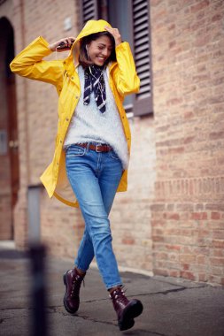 Sarı yağmurluk giyen genç ve yakışıklı bir kadın yağmurlu bir günde şehirde rahat bir şekilde yürürken iyi bir ruh halindedir. Yürü, yağmur, şehir