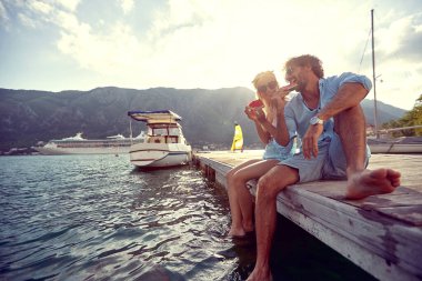 Genç bir çift sahilde rıhtımda oturuyor ve güzel bir gün batımında sulu karpuzun tadını çıkarıyor. Aşk, ilişki, tatil, deniz