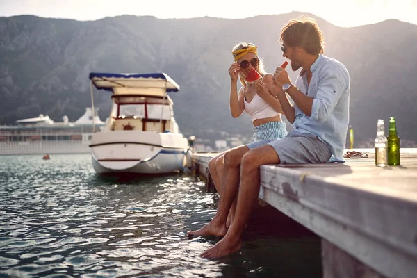 湖の桟橋に座っている笑顔のカップルを愛し 自然を楽しんでスイカを食べる — ストック写真