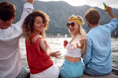 Ahşap iskelede oturan deniz kenarında neşeli genç insanlar. Kızlar ellerini kameraya doğru uzatıyor. Seyahat, turizm, birliktelik, eğlence, yaz tatili konsepti.