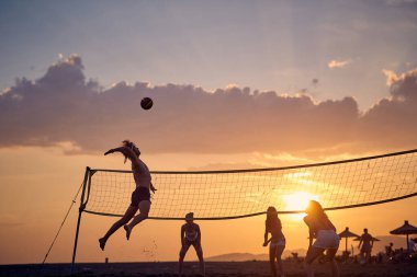 Genç arkadaşlar gün batımında plajda voleybol oynuyorlar.