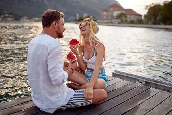 Çiftler deniz kenarında tahta rıhtımda oturup gülerek ve karpuz yiyerek iyi vakit geçiriyorlar. Aşk, eğlence, birliktelik konsepti..