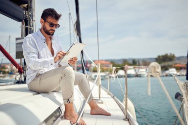 A young businessman is sitting on his yacht and doing some job on his tablet while riding in the dock on a beautiful day on the seaside. Summer, sea, vacation