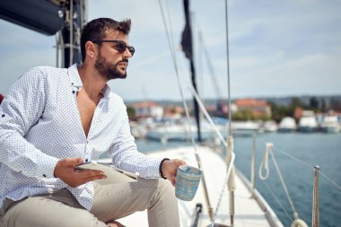 Tabletli genç bir adam, güneşli bir sahil gününde rıhtımın güzel manzarasının ve kahvenin tadını çıkarırken yatta fotoğraf için poz veriyor. Yaz, deniz, tatil