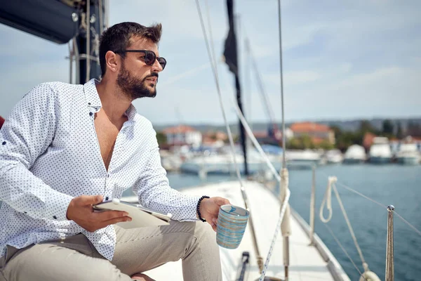 A young man with a tablet is sitting and posing for a photo on the yacht while enjoying a coffee and beautiful scenery of the dock on a sunny day on the seaside. Summer, sea, vacation