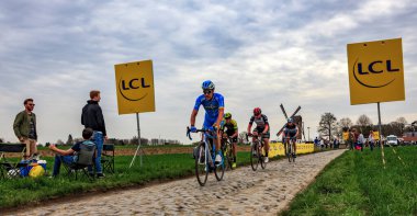 Templeuve, Fransa - 08 Nisan 2018: Paris-Roubaix 2018 sırasında Templeuve geleneksel Vertain yel değirmeni önünde Arnavut kaldırımlı yolda sürme peloton. 