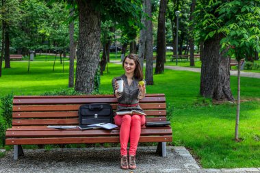 Parkta ders çalışan genç bir kadın elinde yeşil bir elma ve bir fincan kahveyle kameraya bakıyor.. 
