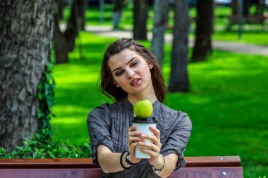 Parkta ders çalışan genç bir kadın elinde yeşil bir elma ve bir fincan kahveyle kameraya bakıyor.. 