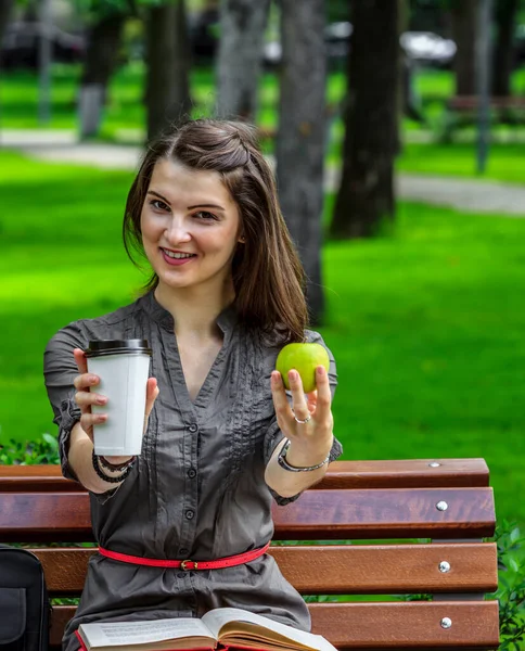 一个在公园里学习的年轻女人拿着一个绿色的苹果和一杯咖啡 望着摄像机 — 图库照片